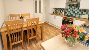 A compact kitchen with white cabinets, a wooden dining table with chairs, a small stove, and a vase with flowers on the counter. - Station View (Whitby)