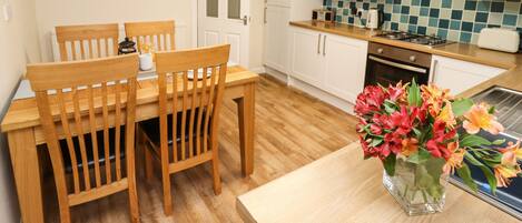 A compact kitchen with white cabinets, a wooden dining table with chairs, a small stove, and a vase with flowers on the counter.