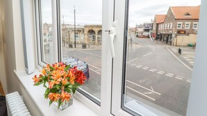 A window with a view of a street and buildings, a vase with flowers on the windowsill, and a radiator below. - Station View (Whitby)