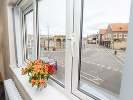 Una ventana con vista a una calle y edificios, un florero con flores en el alféizar, y un radiador debajo.