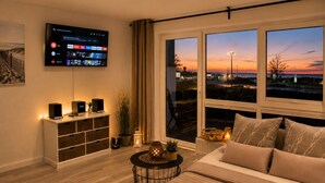 A modern living room with a large TV, a sofa, a coffee table, and a view of the sunset through large windows. - Beautiful apartment directly on the beach Living room and terrace with panoramic sea view     (Heiligenhafen)