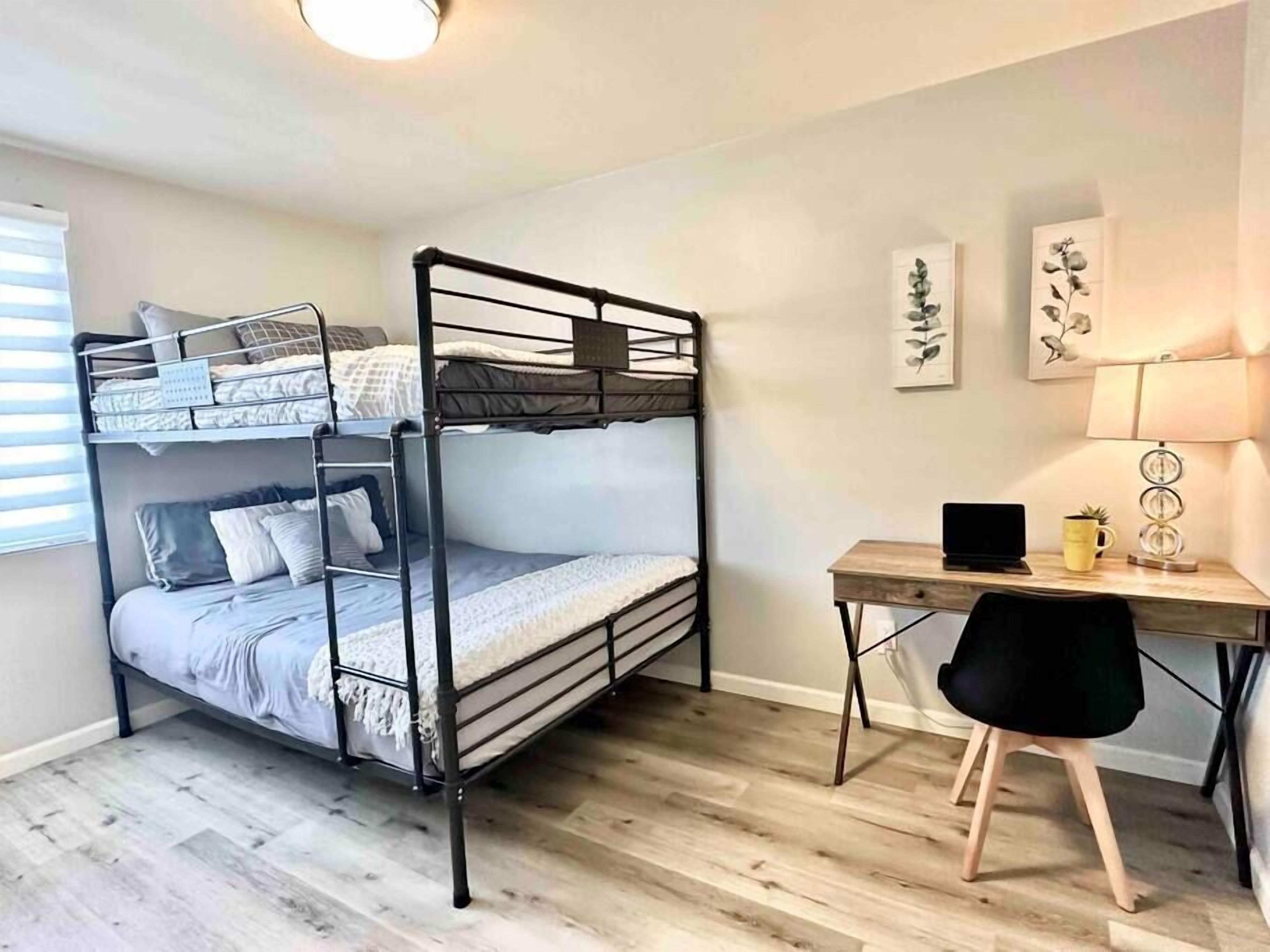 En køjeseng med skrivebord og stol, en lampe samt billedkunst på væggen i et rum med trægulv.