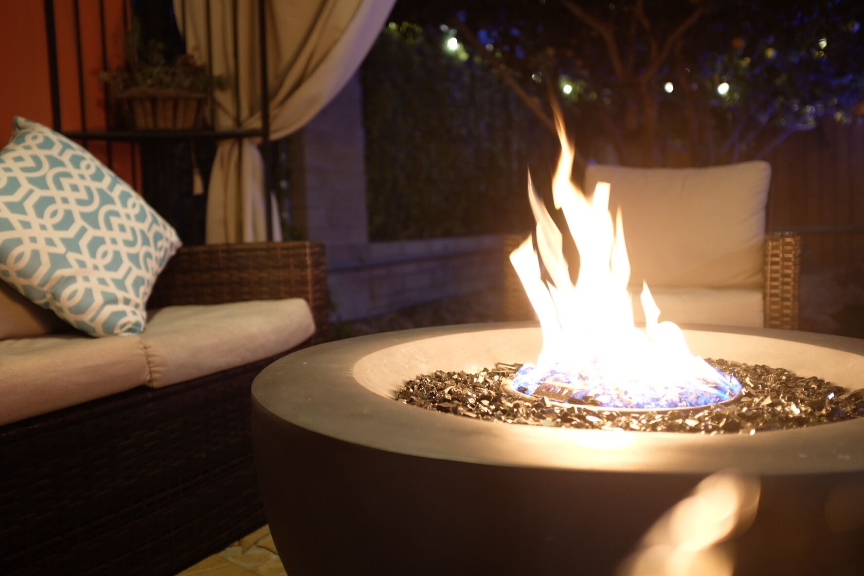 A round outdoor fire feature with a visible flame, surrounded by a dark-colored base, set on a patio with a cushioned seating area and a patterned pillow.