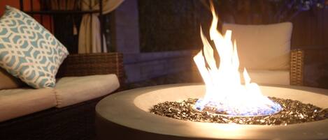 A round outdoor fire feature with a visible flame, surrounded by a dark-colored base, set on a patio with a cushioned seating area and a patterned pillow.