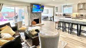 A spacious living room with a fireplace, a large flat-screen TV mounted on a stone wall, a dining area with bar stools, and a kitchen with modern appliances. - Prime Location: Lux 4BR w Pool/Spa/Cabana/Firepit (San Diego)