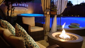 A patio area with a wicker sofa, a round table with a fire pit, and a pool in the background. - Prime Location: Lux 4BR w Pool/Spa/Cabana/Firepit (San Diego)