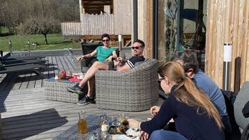 Un grupo de personas descansando en muebles de mimbre al aire libre sobre una terraza de madera, con un edificio moderno al fondo.