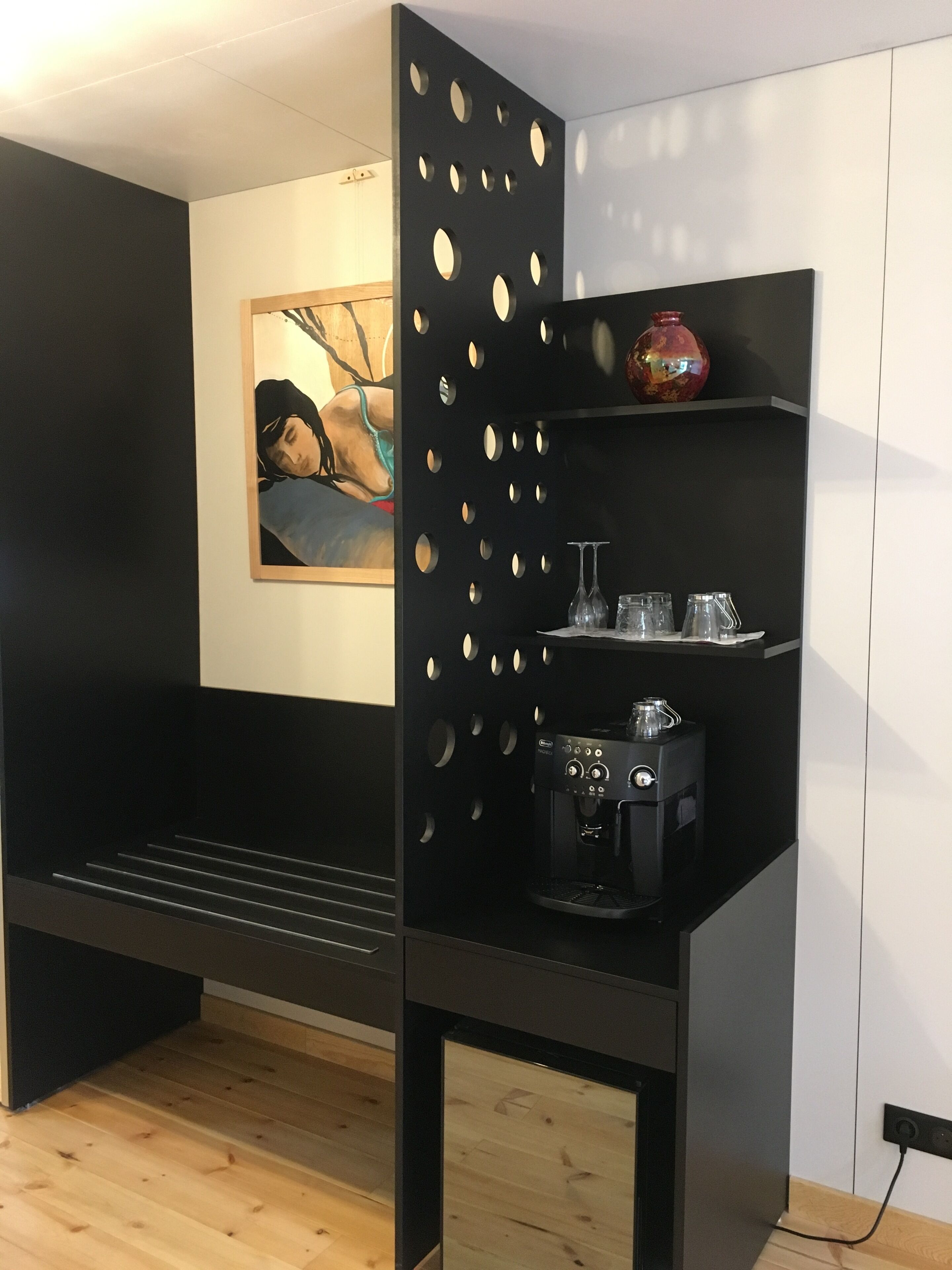 A modern room with a black partition featuring circular cutouts, a coffee machine, and a shelf with decorative items.