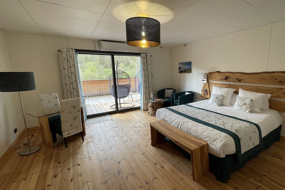 A bedroom with a large bed, a wooden headboard, a bench, a desk, a chair, and a view through a glass door to an outdoor area.