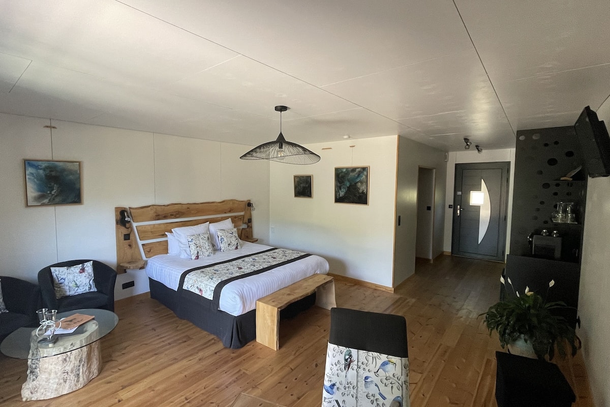 A bedroom with a wooden bed, a small bedside table, a chair, and a bench. There is a door leading to a kitchenette and a bathroom.