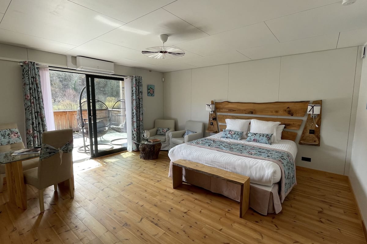 A bedroom with a wooden floor, a bed with a wooden headboard, a desk with a chair, and a view through a glass door.