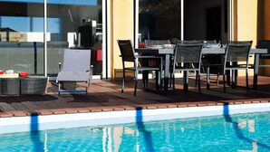 An outdoor pool area with a table and chairs, a covered seating area, and a glass door leading to a building. - Family villa with pool – 6 guests (Les Sables-d´Olonne)