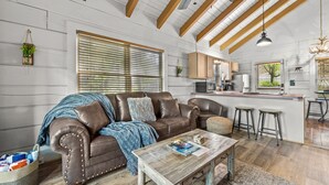 A spacious living room with a brown leather sofa, wooden coffee table, and a kitchen area with wooden cabinets and a bar stool. - Rustic Retreat in the Smokies (Sevierville)