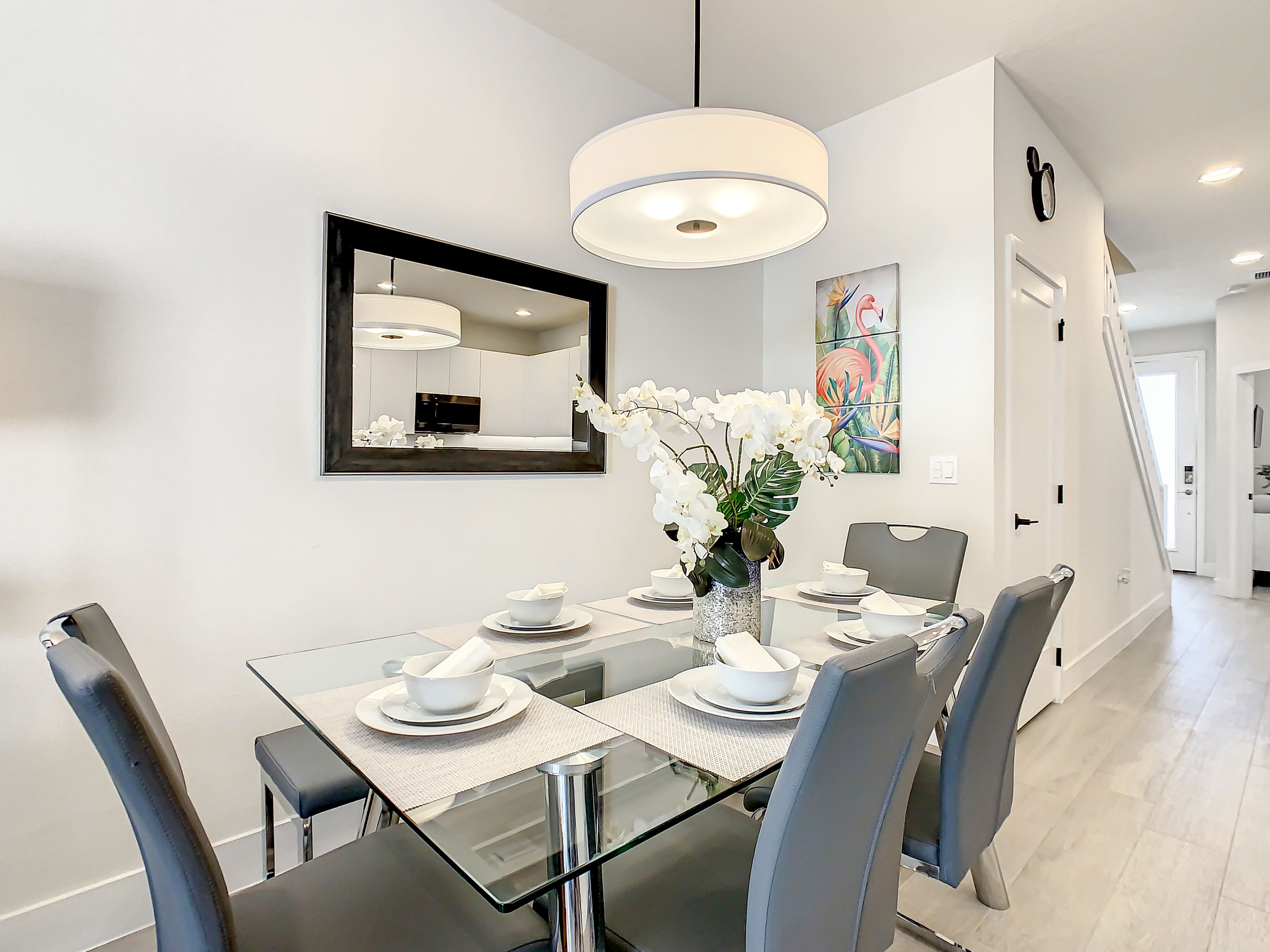 Een moderne eetruimte met een glazen tafel, grijze stoelen, een witte vaas met bloemen en een grote spiegel aan de muur.