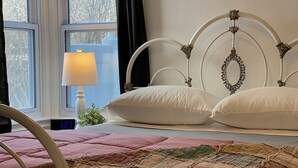 Een bed met een metalen frame, witte kussens en een sprei. Er staat een nachtkastje met een lamp en een wekker.