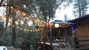 Une cabane en bois rustique avec une terrasse, entourée d’arbres et illuminée de guirlandes lumineuses.