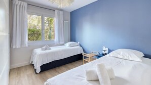 Ein Zimmer mit zwei Betten, einem fensterartigen Ausblick mit Vorhängen, einer weißen Deckenleuchte und Blick ins Grüne.
