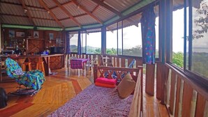 Een ruime kamer met een houten vloer, een bank met kussens, een kleurrijke fauteuil, een eettafel met stoelen en grote ramen met uitzicht op het landschap.