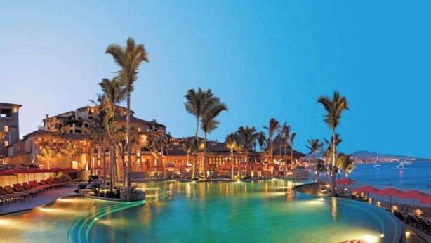 Un resort con una piscina grande, palmeras y un edificio con varios balcones con vista al agua.