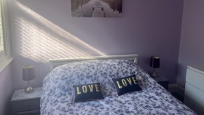 A bedroom with a bed, a nightstand, a lamp, a wall-mounted picture, and a window with sunlight. - Spacious 1-bed Apartment in the Heart of Herne Bay (Herne Bay)