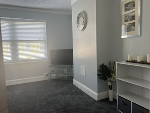 A room with a grey carpet, a desk with a flat-screen TV, a clock on the wall, and a shelf with candles.