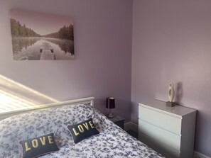 A bedroom with a bed, a nightstand, a dresser, a lamp, and a painting of a lake.