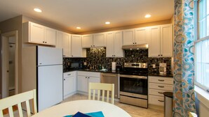 A modern kitchen with white cabinets, a black countertop, and stainless steel appliances. There is a dining table with chairs and a book on it. - Heart of Downtown Bar Harbor - fully airconditioned (Bar Harbor)
