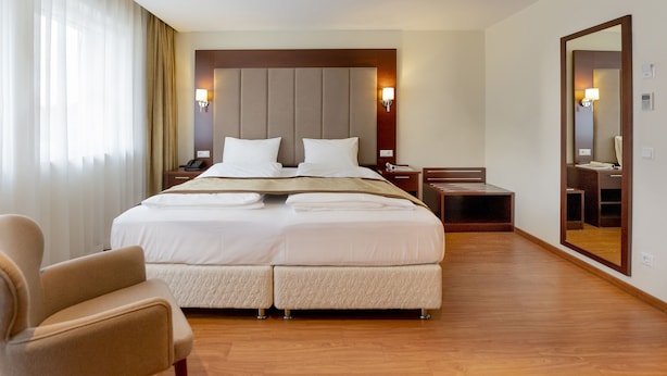 Una habitación de hotel con una cama grande, una silla, una mesita de noche, un espejo y una ventana con cortinas.