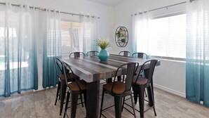 A dining area with a wooden table and chairs, a vase with a plant, and a wall-mounted shelf with books and a plant.