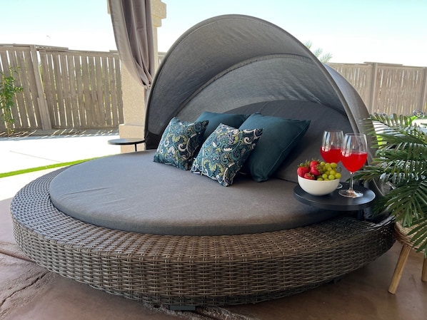 A round outdoor seating area with a cushioned bench, two patterned pillows, a bowl of fruit, and two glasses of red drink.