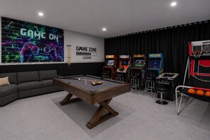Ein Billardtisch, eine Couch und Spielautomaten in einem Raum mit dem Schriftzug 'GAME ON'.