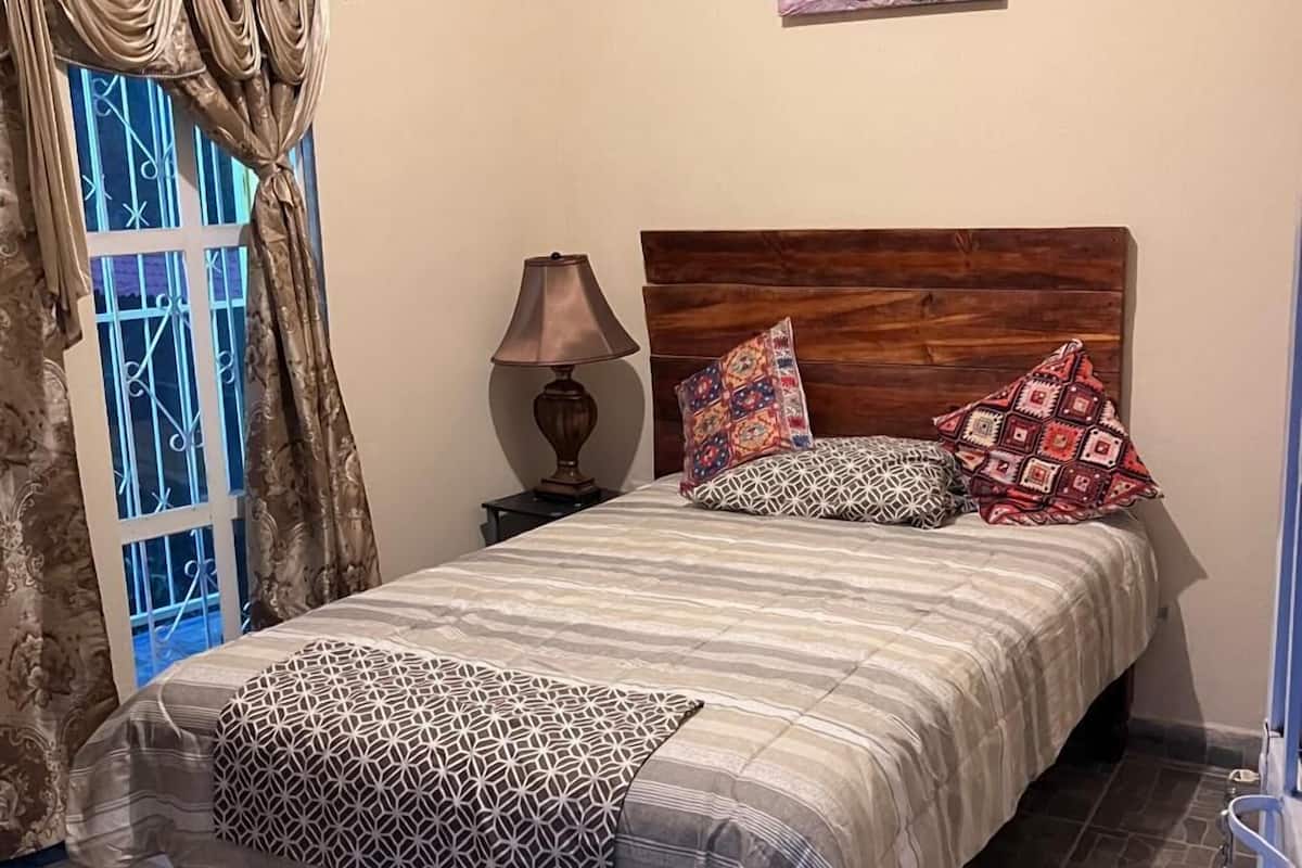 A bedroom with a bed, a wooden headboard, patterned pillows, a nightstand with a lamp, a window with curtains, and a painting on the wall.