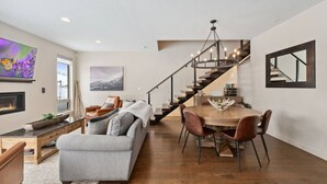 A modern living room with a fireplace, a dining area with a wooden table and chairs, a staircase, and a large painting on the wall.