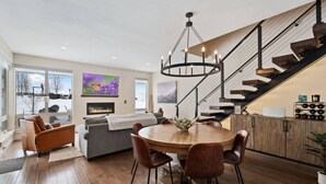 A modern living room with a fireplace, a dining area with a round table and chairs, and a staircase with a black railing.