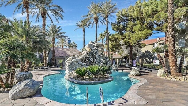 A pool area with a central rock feature, surrounded by palm trees and a stone pathway. - Vegas Resort 2-Bed Condo walking distance to Casinos (Ideal for Long Stays) (Las Vegas)
