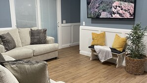 A living room with a beige sofa, a white bench, and a large flat-screen TV mounted on the wall. - Vegas Resort 2-Bed Condo walking distance to Casinos (Ideal for Long Stays) (Las Vegas)