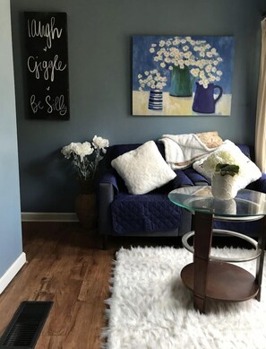 Un salon avec un canapé bleu foncé, une table basse en verre, un tapis blanc et moelleux, et un tableau représentant des fleurs accroché au mur.