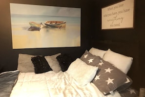 Une chambre à coucher avec un lit, des oreillers, un tableau représentant des bateaux accroché au mur, et un panneau affichant une citation inspirante.
