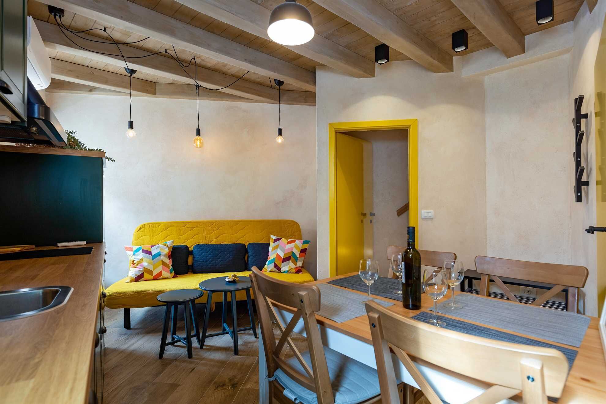 Un salon chaleureux avec un canapé jaune, une table à manger en bois et des chaises. On peut apercevoir un téléviseur et un petit coin cuisine.