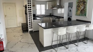 A modern kitchen with a central island, white bar stools, and a large window providing natural light. - Nice villa in La Cala de Mijas  Spain with heated pool and  sea ​​view. (Las Lagunas de Mijas)