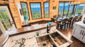 A wooden interior with a kitchen and dining area, large windows with mountain views, and a sink with a faucet.