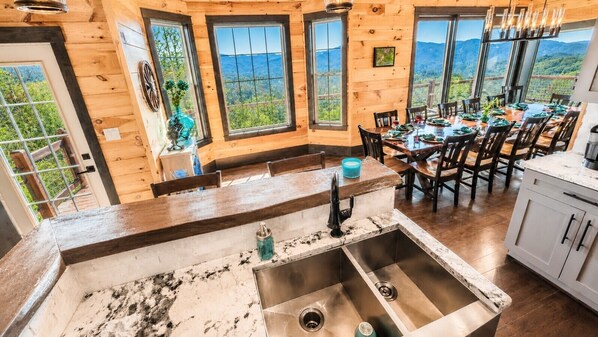 Een houten interieur met een keuken en een eetgedeelte, grote ramen met uitzicht op de bergen, en een spoelbak met een kraan.