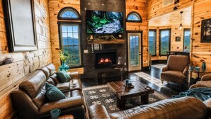 A cozy cabin interior with a fireplace, leather seating, a television, and a scenic view.