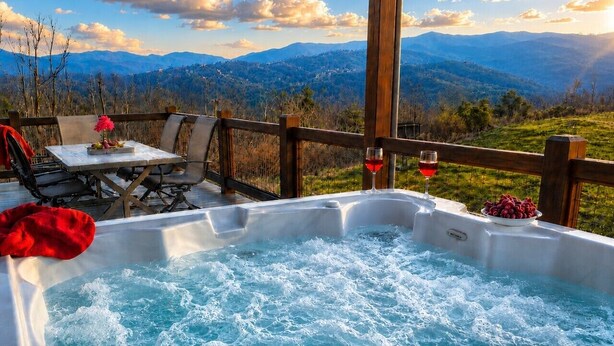 A hot tub with a mountain view, a table with chairs, and a glass of red wine.