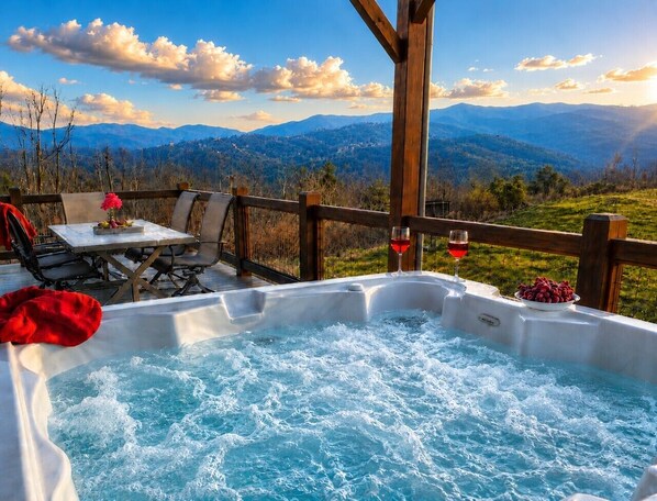 A hot tub with a mountain view, a table with chairs, and a glass of red wine.
