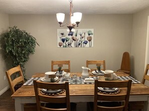 A dining room with a wooden table set for four, surrounded by wooden chairs. There is a chandelier above the table, a potted plant to the left, and a floral painting on the wall.