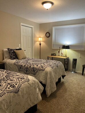 A bedroom with two beds, a bedside table, a lamp, a heater, a clock, and a window with blinds.