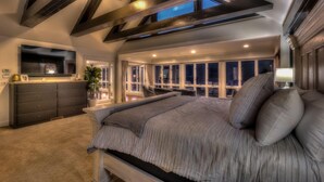 Ein Schlafzimmer mit einem großen Bett, einem Schminktisch, einem Fernseher und Blick auf den Nachthimmel durch die Fenster.