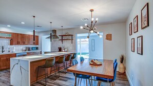 A modern kitchen with wooden cabinets, a central island, and a dining area with a wooden table and blue chairs. - South Scottsdale Poolside Getaway! (Scottsdale)