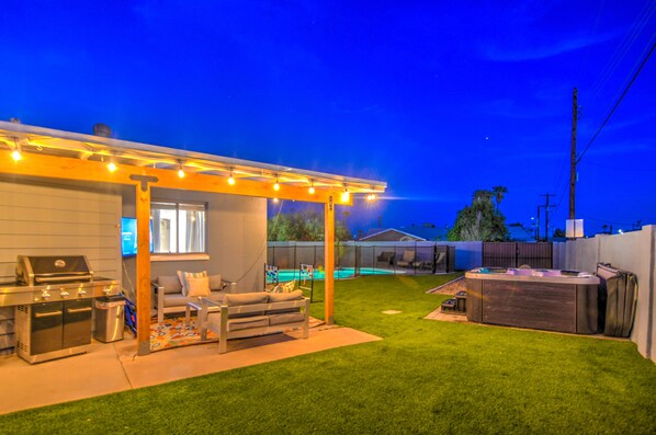 Relax in the backyard with a covered seating area, barbecue grill, and hot tub, surrounded by a lush lawn and a pool.